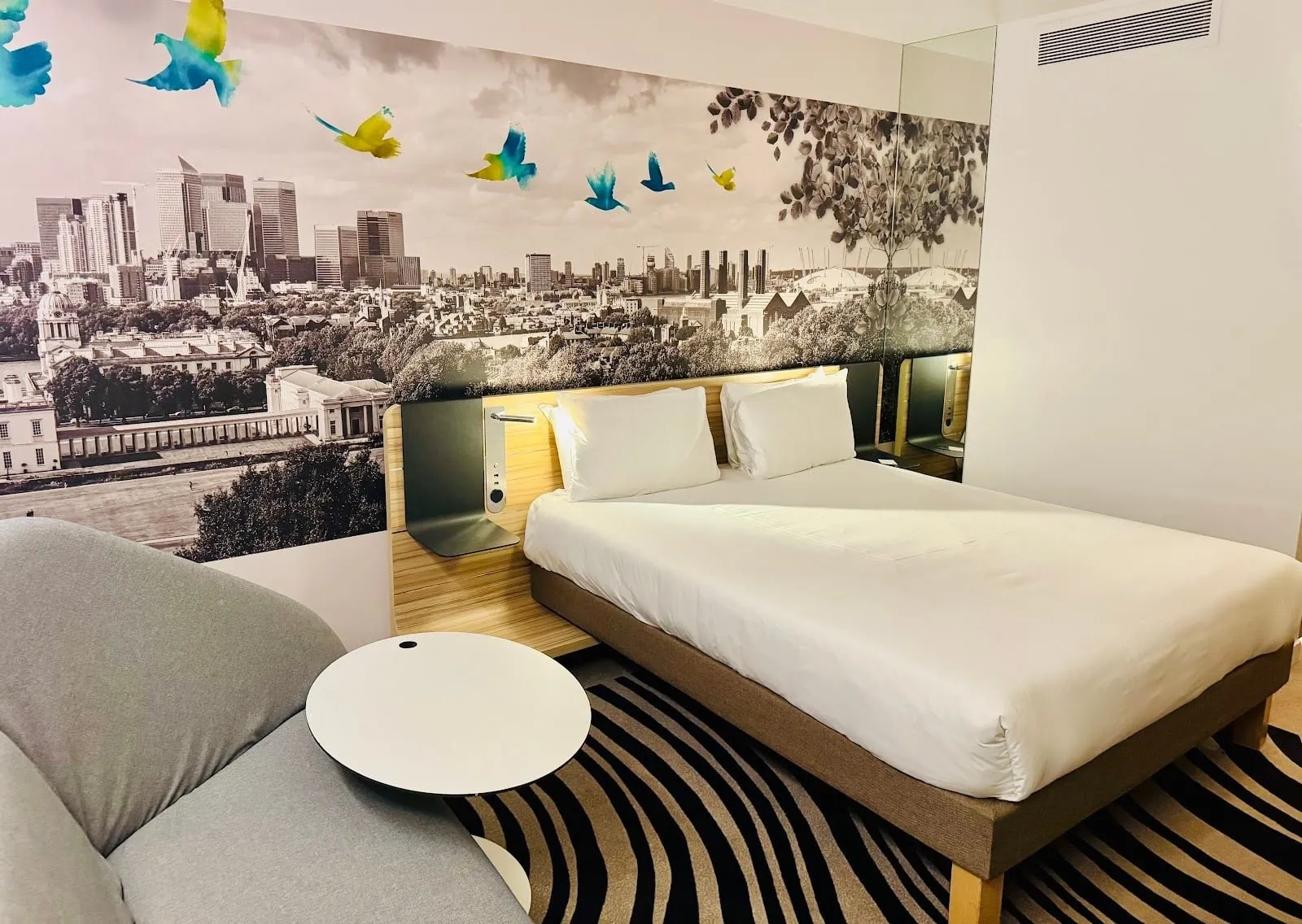 A room at Novotel London ExCeL with a double bed, a grey sofa and a mural of the Greenwich and Canary Wharf skyline