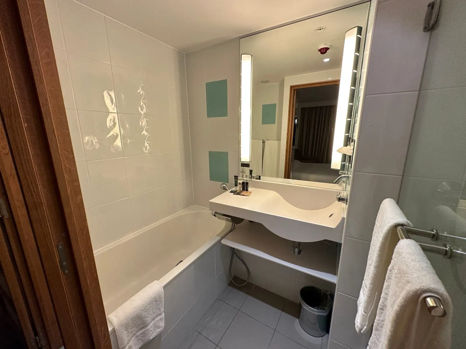 A white-tiled bathroom at Novotel London Paddington with a bath, a sculpted basin under a lit mirror and a towel rail beside a glass screen