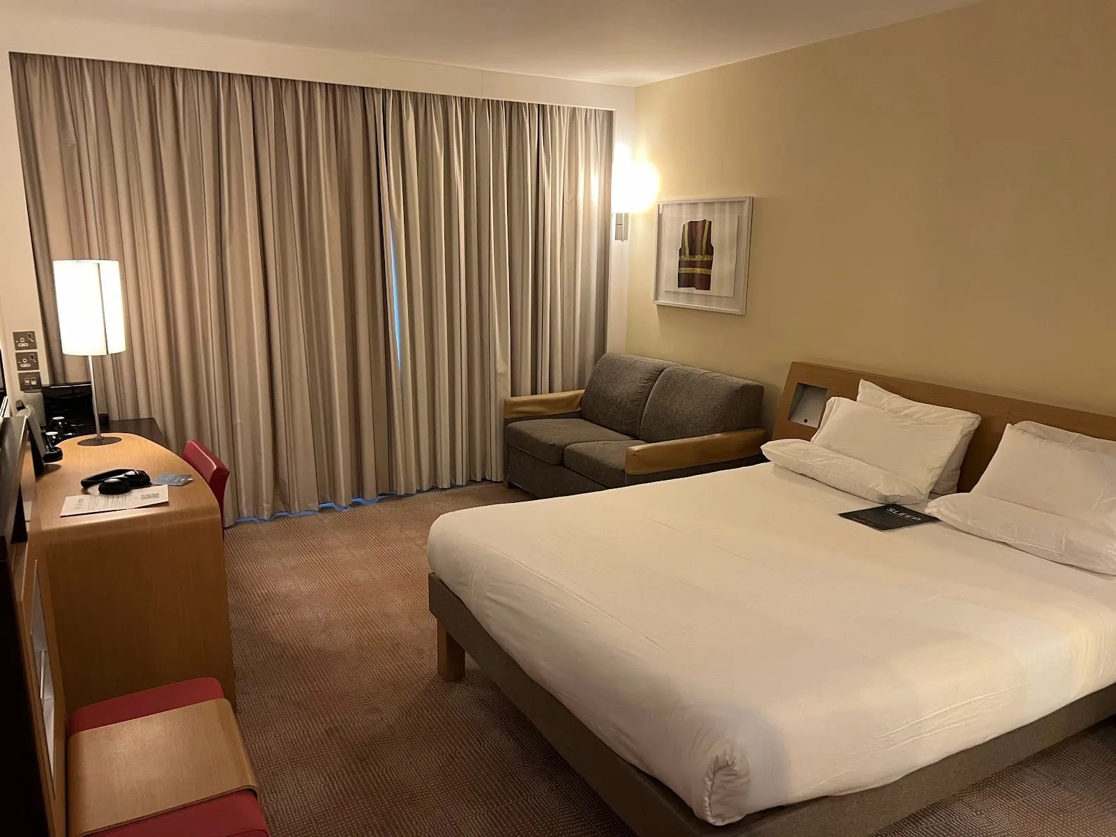 A room at Novotel London Paddington with a queen bed on a light wood frame, a grey two-seat sofa under floor-length curtains, and a curved wood desk with a red chair