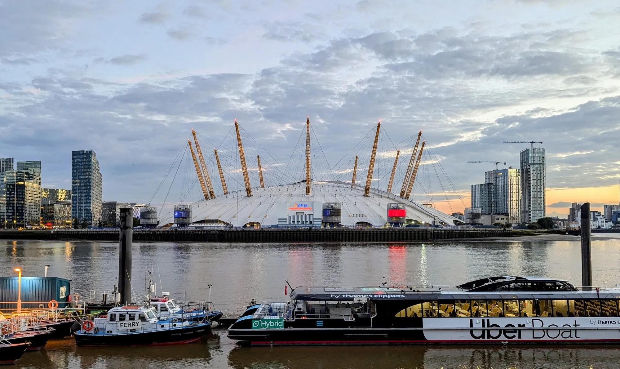 The O2's white dome and yellow support masts seen from across the Thames at dusk, with an Uber Boat by Thames Clippers moored in front