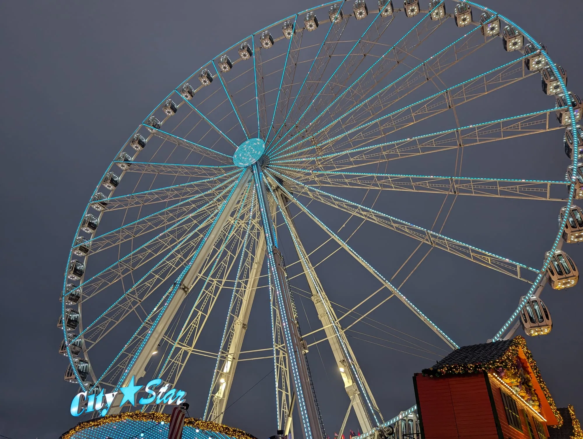 The observation wheel at Winter Wonderland lit up against a dusk sky, its spokes picked out in turquoise lights, with an illuminated stall roof below
