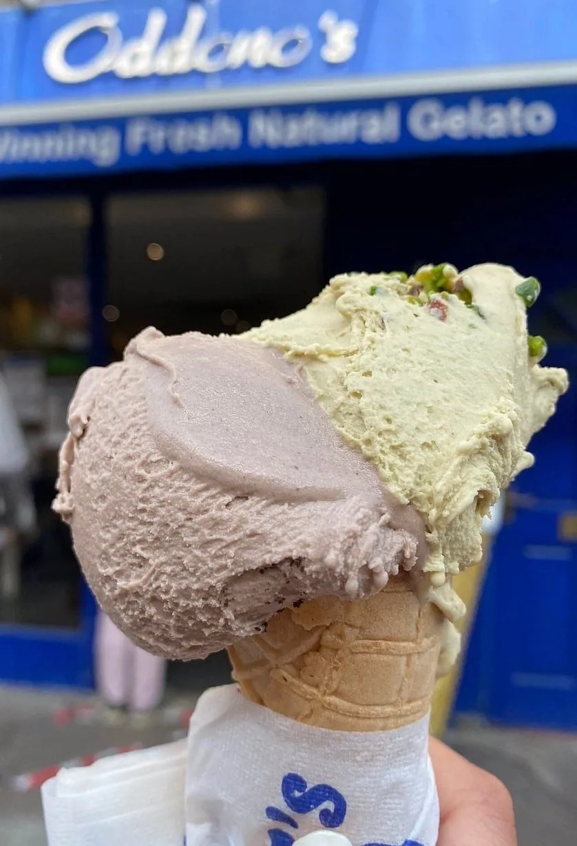 A double cone held up in front of the blue Oddono's shopfront, its sign reading Fresh Natural Gelato