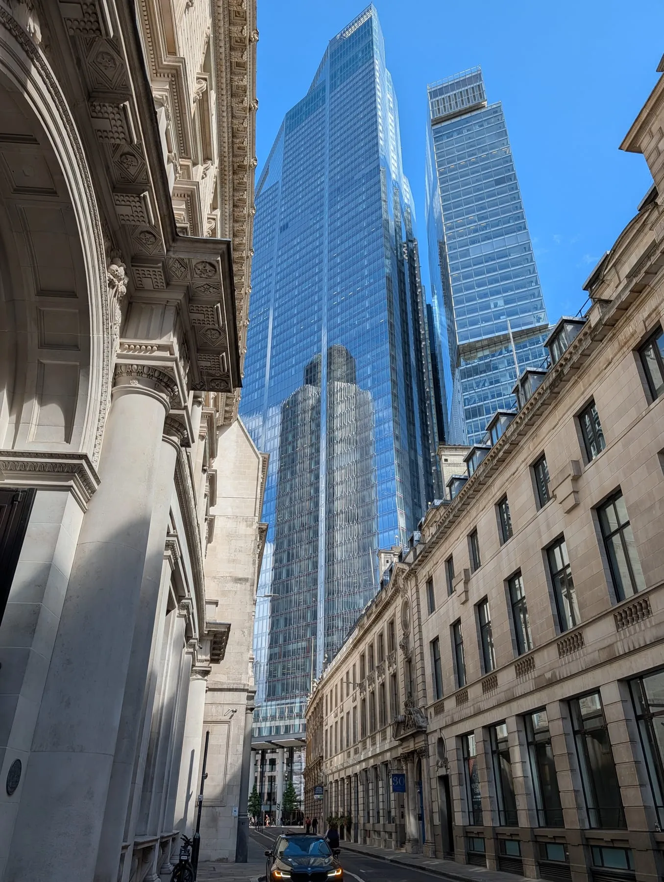 A classical stone portico and carved cornice in the foreground with the glass towers of 22 Bishopsgate and the Scalpel rising directly behind