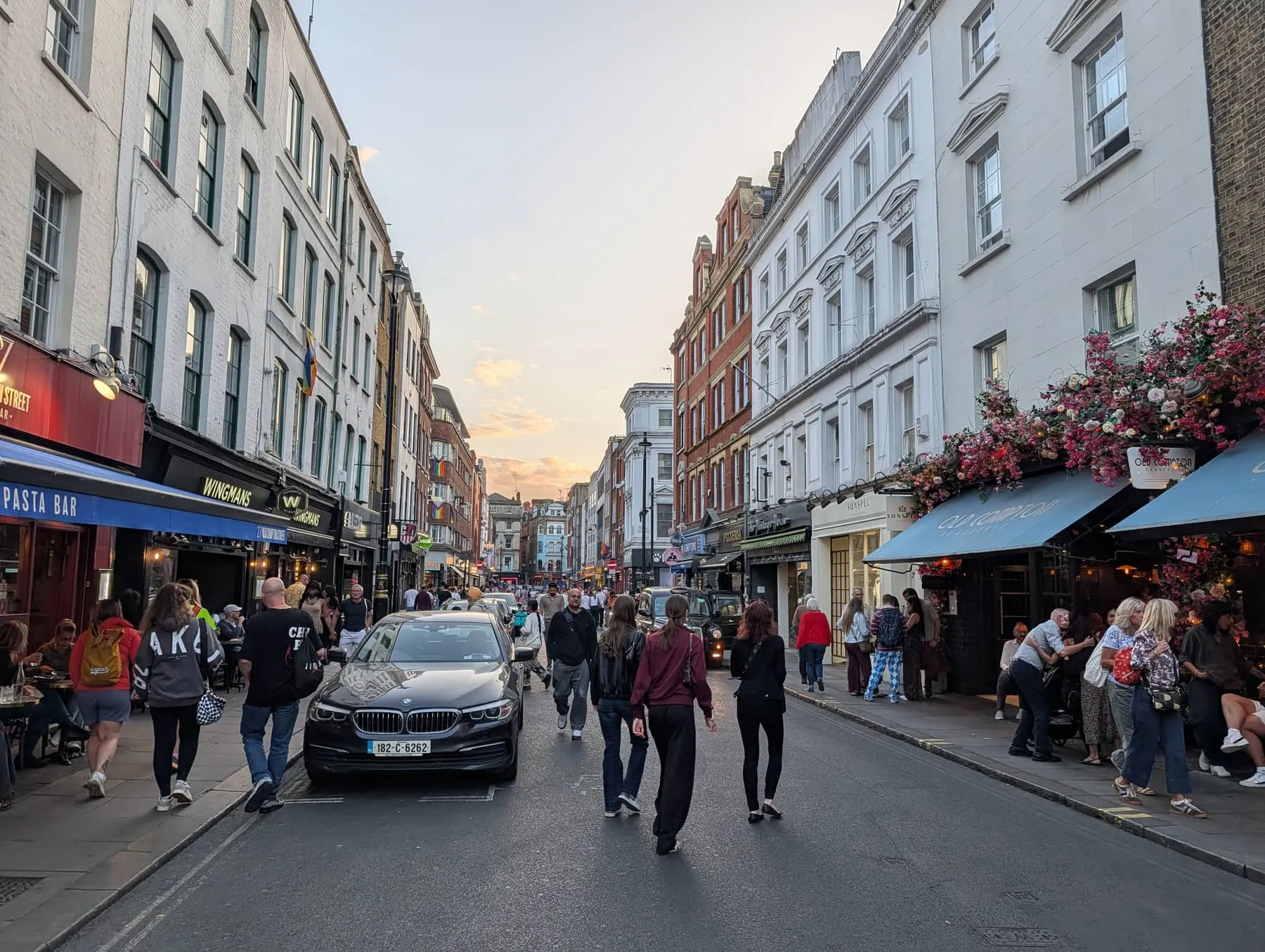 Old Compton Street at dusk, busy with people, lined with bars and a florally decorated frontage