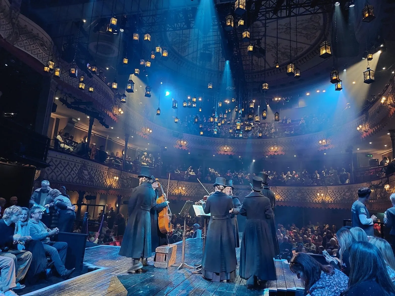 Hundreds of lit lanterns hanging above the audience at the Old Vic, with musicians in top hats and long coats playing on a wooden stage between the seats