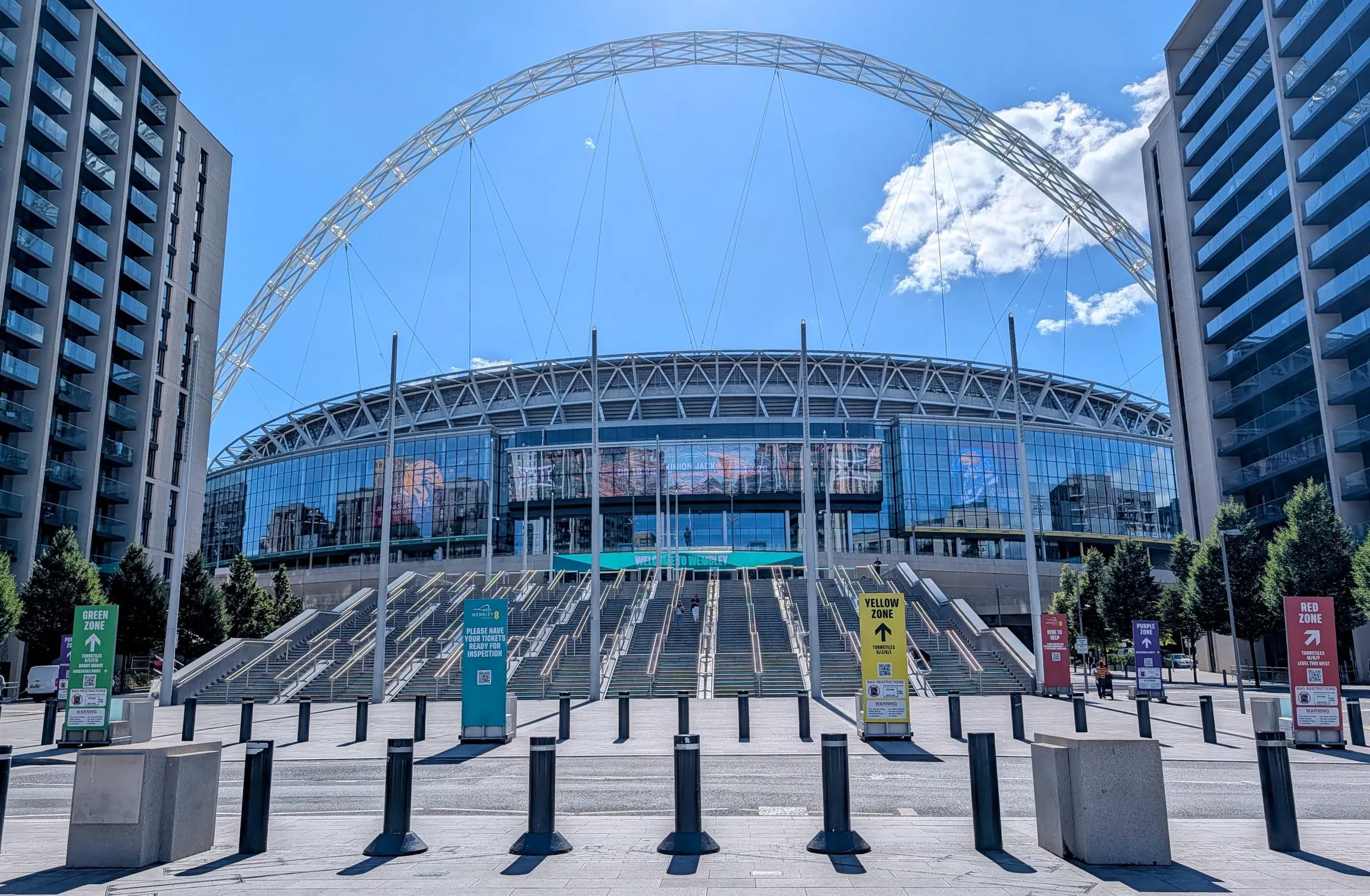 Wembley Stadium's glass front and arch seen from the foot of the Olympic Steps, bollards across the plaza in the foreground