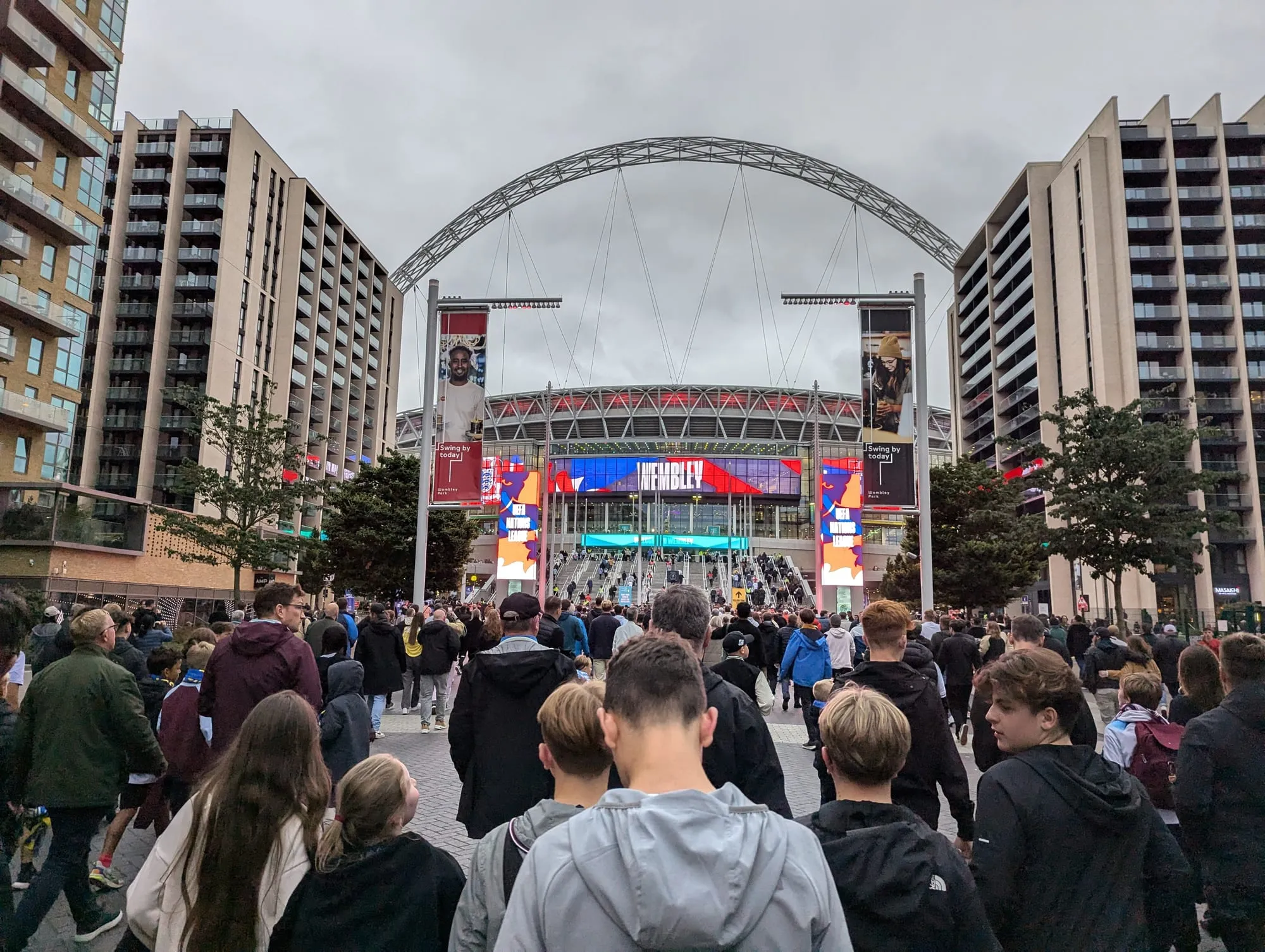 A crowd walking up Olympic Way towards Wembley Stadium before kick-off, the arch above the roof and the stadium screens lit ahead