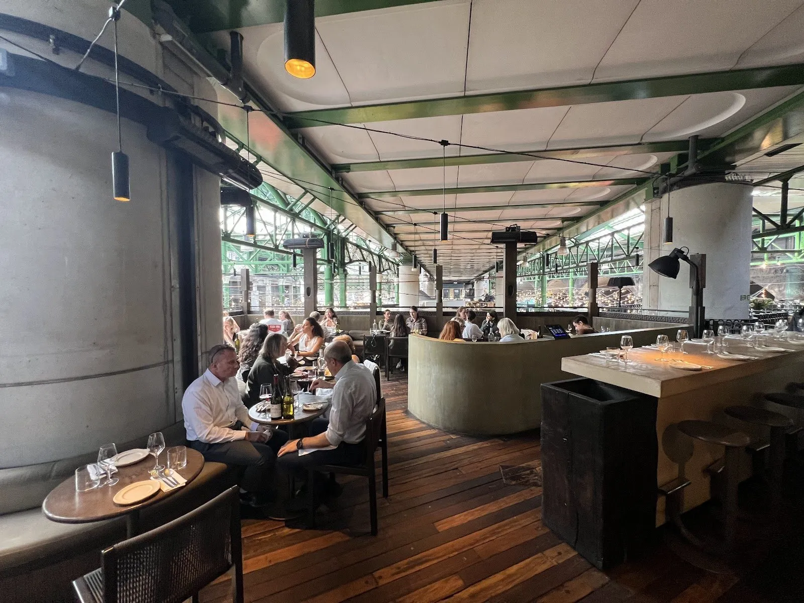 Oma's dining room under the green iron roof of Borough Market, with diners at round tables on a wooden floor, a curved concrete counter and pendant lights