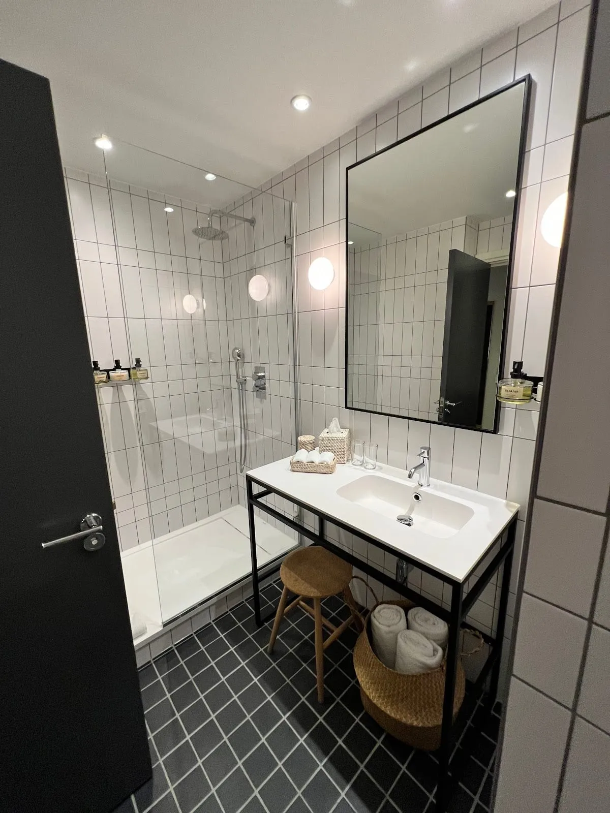 A bathroom at One Hundred Shoreditch with white subway tiles, a black-framed mirror and a walk-in glass shower, with rolled towels in a wicker basket
