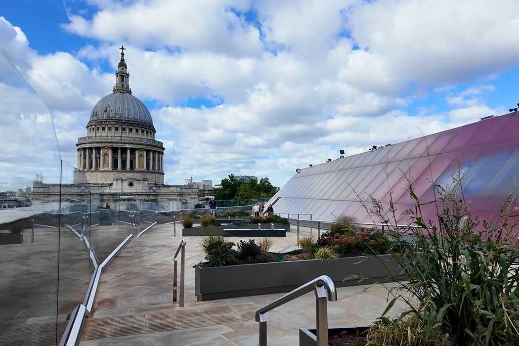 The dome of St Paul's Cathedral behind a glass balustrade, with planted beds on a paved roof terrace and a tilted glass roof to the right under a blue sky