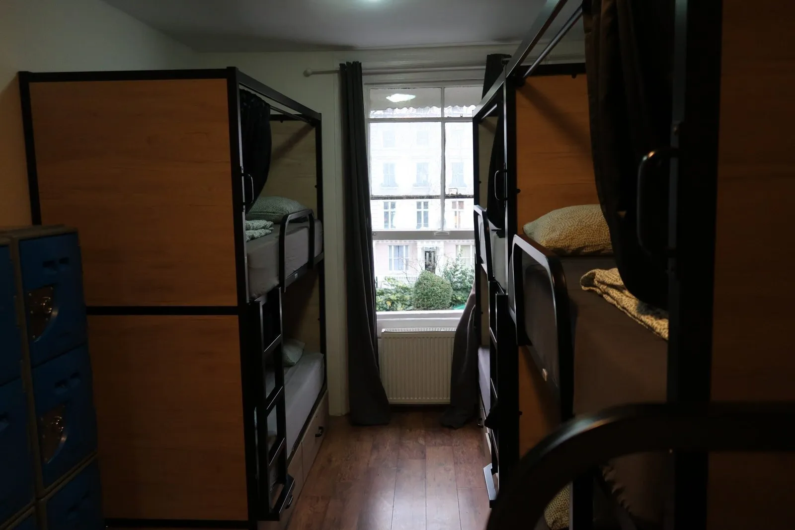 A dorm at Onefam Notting Hill with wood-panelled bunk beds with dark curtains on each side, blue lockers by the door and a sash window onto the stucco houses of Prince's Square