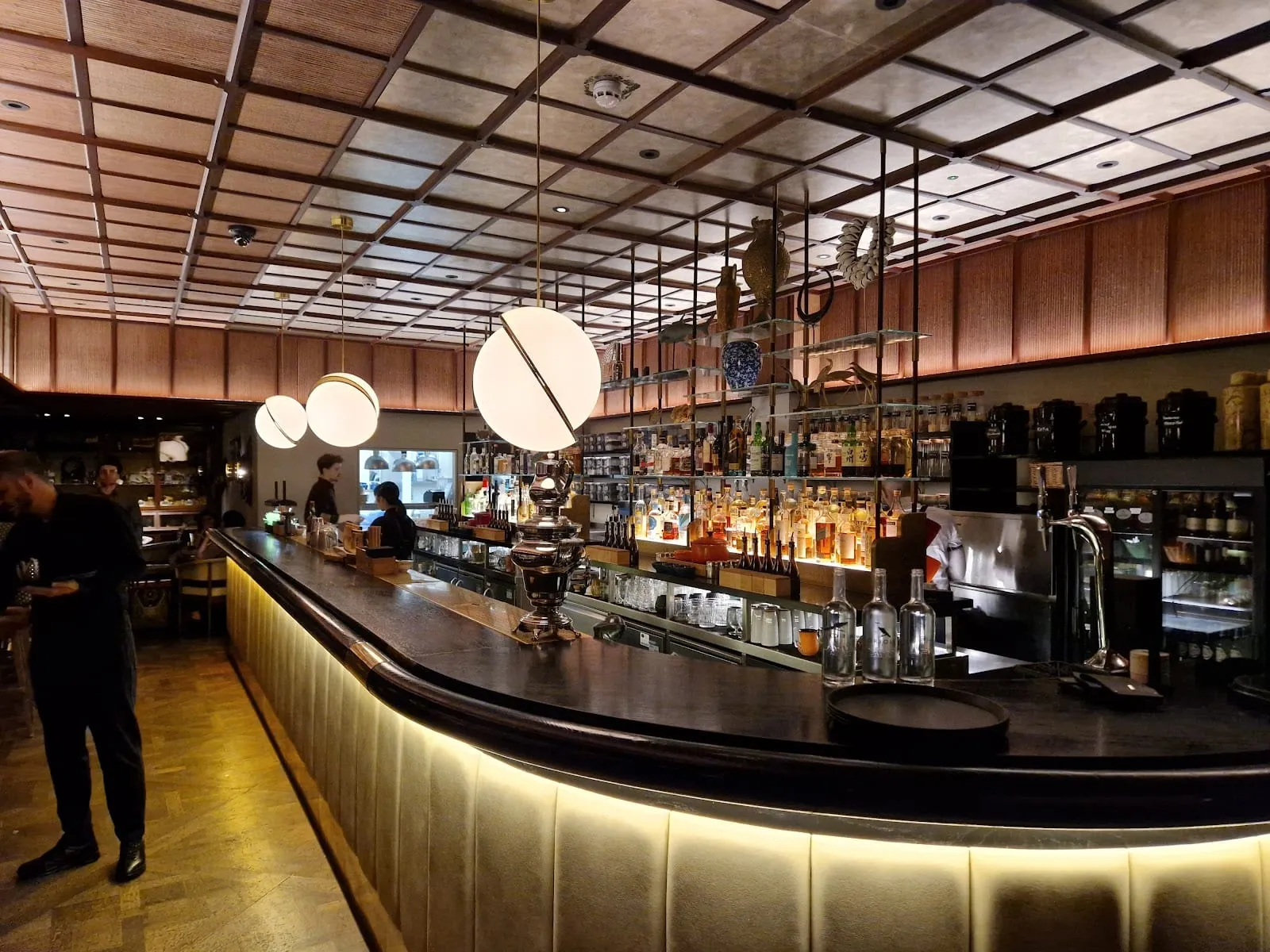 A long curved bar with a lit upholstered front and a dark wood top, under globe pendant lights and a coffered ceiling, with glass shelves of bottles and ornaments behind