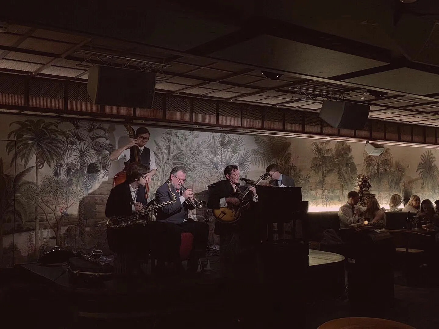 A five-piece jazz band plays in front of a palm-tree mural at Oriole, with diners at a booth to one side