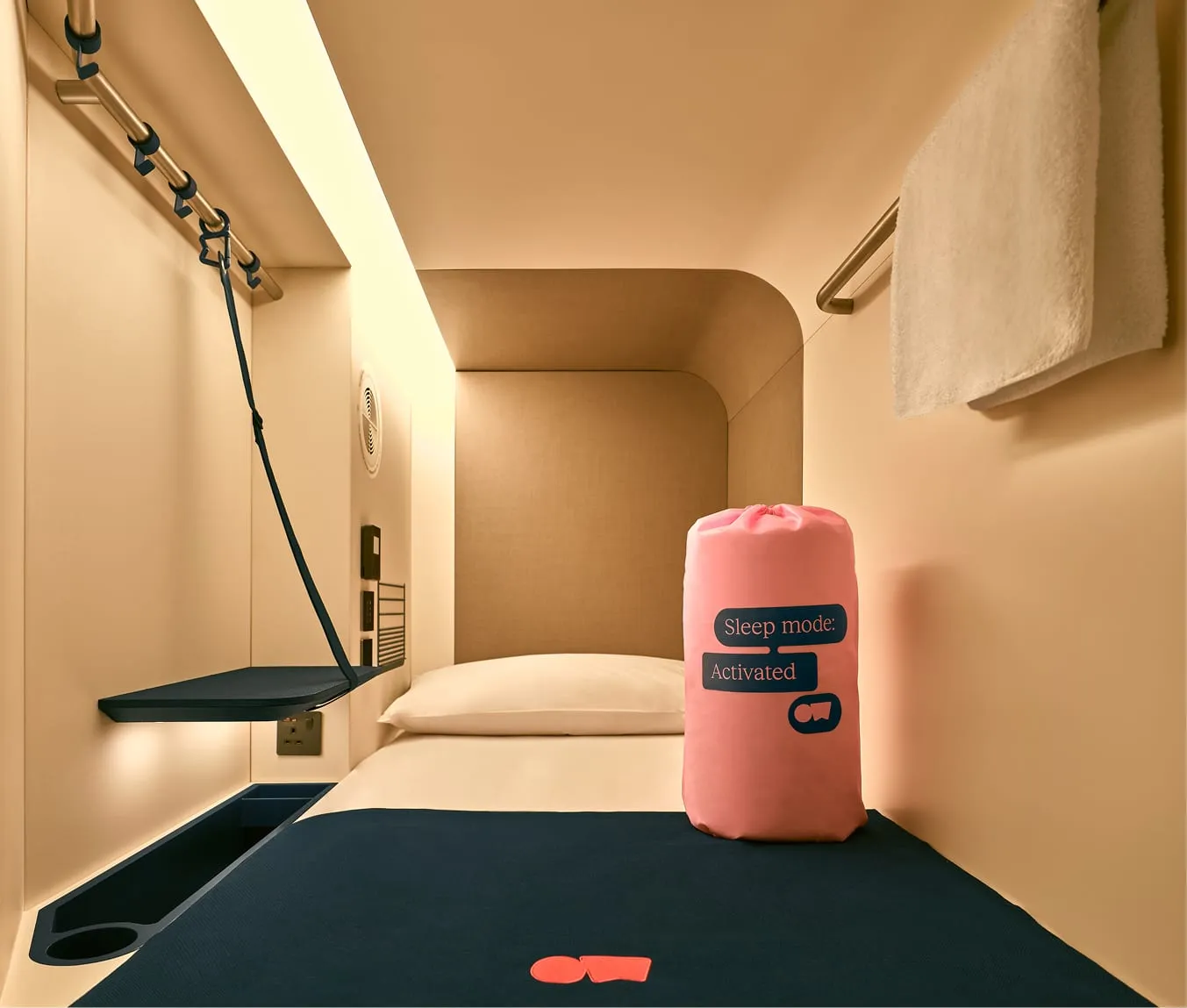 Inside an Otherwander single nest: a cream-lined pod with a fold-down shelf, hanging rail and hooks, a UK socket, an air vent, a towel rail and a pink bag reading Sleep mode: Activated