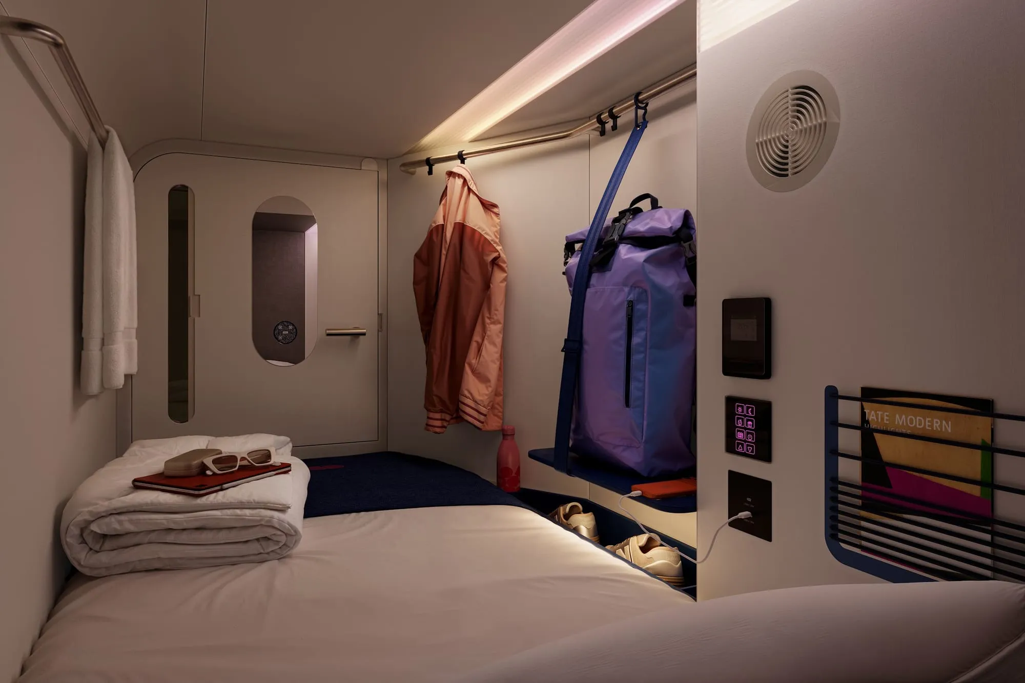 An Otherwander Soho sleeping pod with a folded duvet, an orange jacket and a purple backpack hanging beside it, and a bedside touchscreen control panel