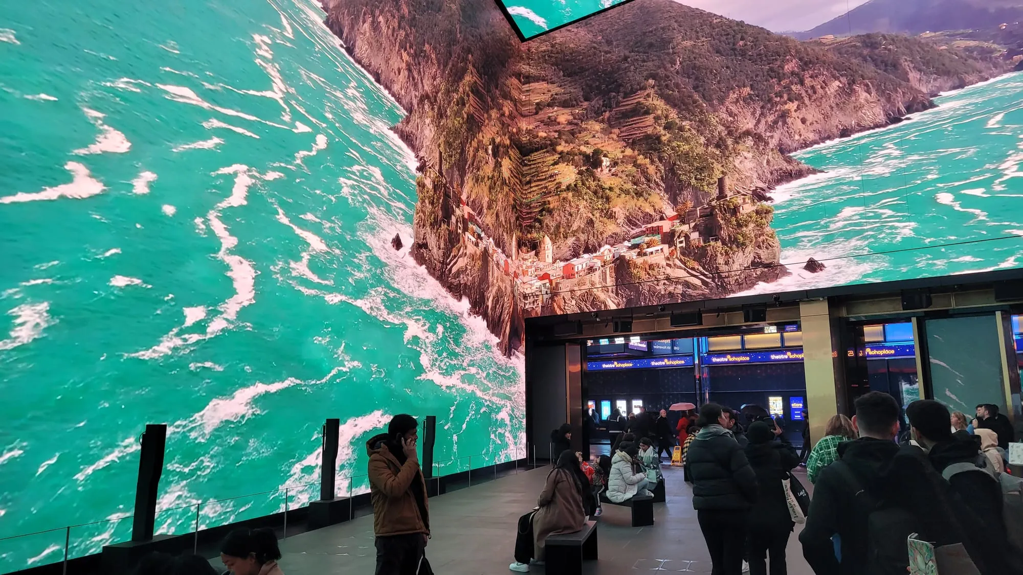 The wraparound screens of the Now Building at Outernet showing a coastline from above, with people watching from the floor below