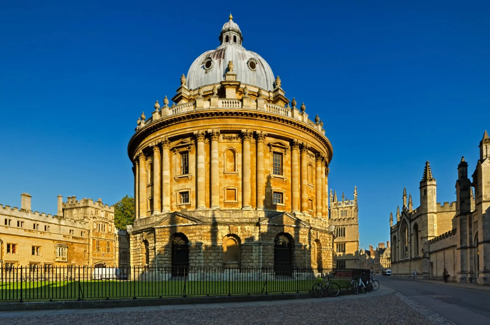 The round, domed Radcliffe Camera in honey-coloured stone, behind iron railings and a lawn, with college buildings either side under a deep blue sky