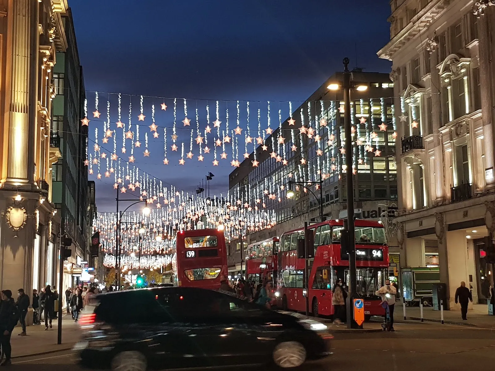 Rows of white stars on hanging strands of light strung across Oxford Street at dusk, with red buses at Oxford Circus below
