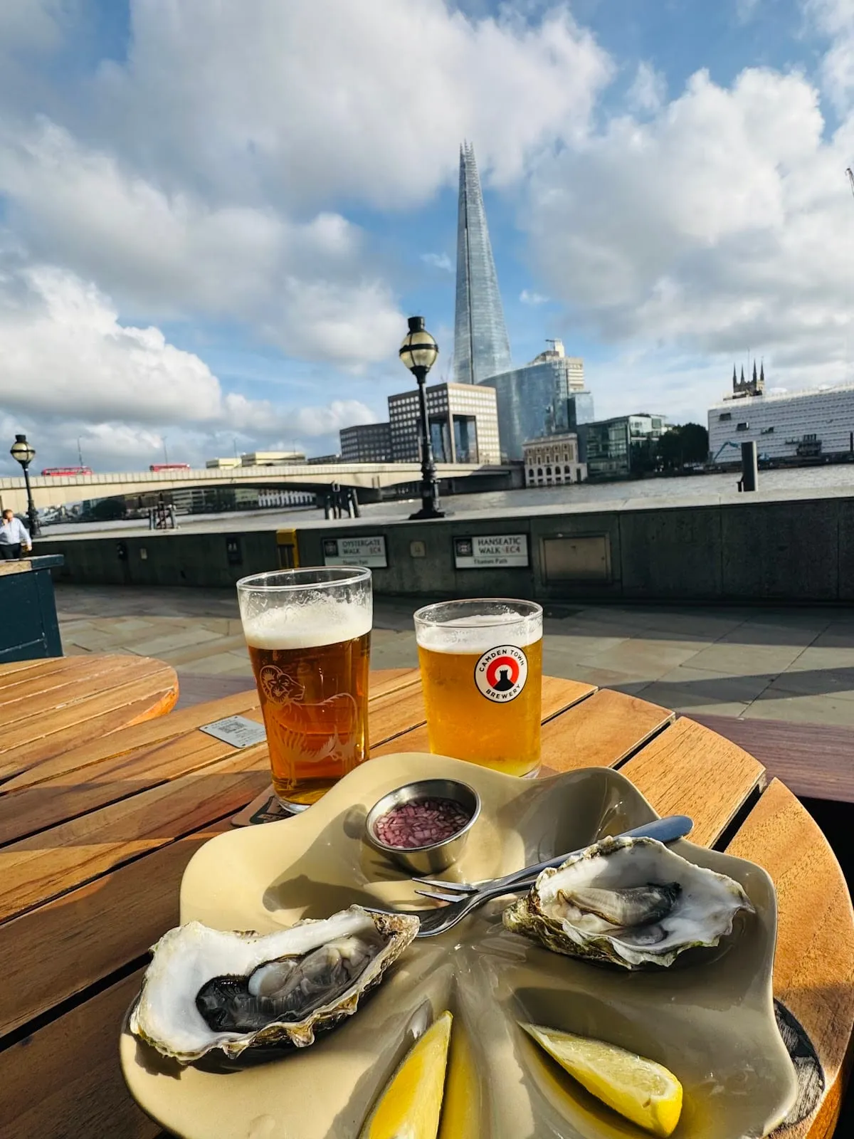 A plate of oysters with lemon and two pints on an outdoor table by the Thames, with London Bridge and the Shard across the river
