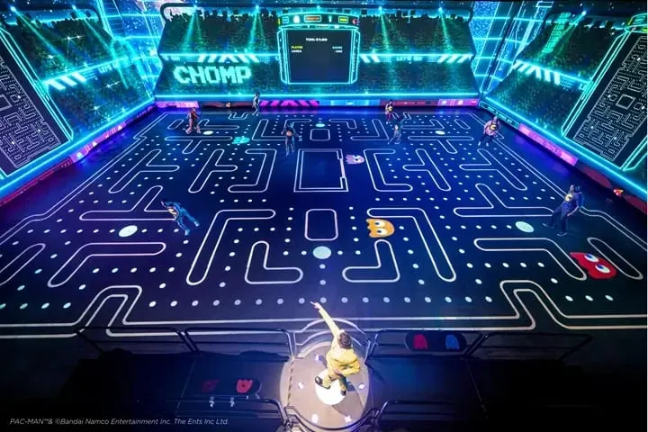 A vast illuminated maze laid out across a dark floor in glowing blue and white lines and dots, with players running the corridors and a scoreboard reading CHOMP on the wall above