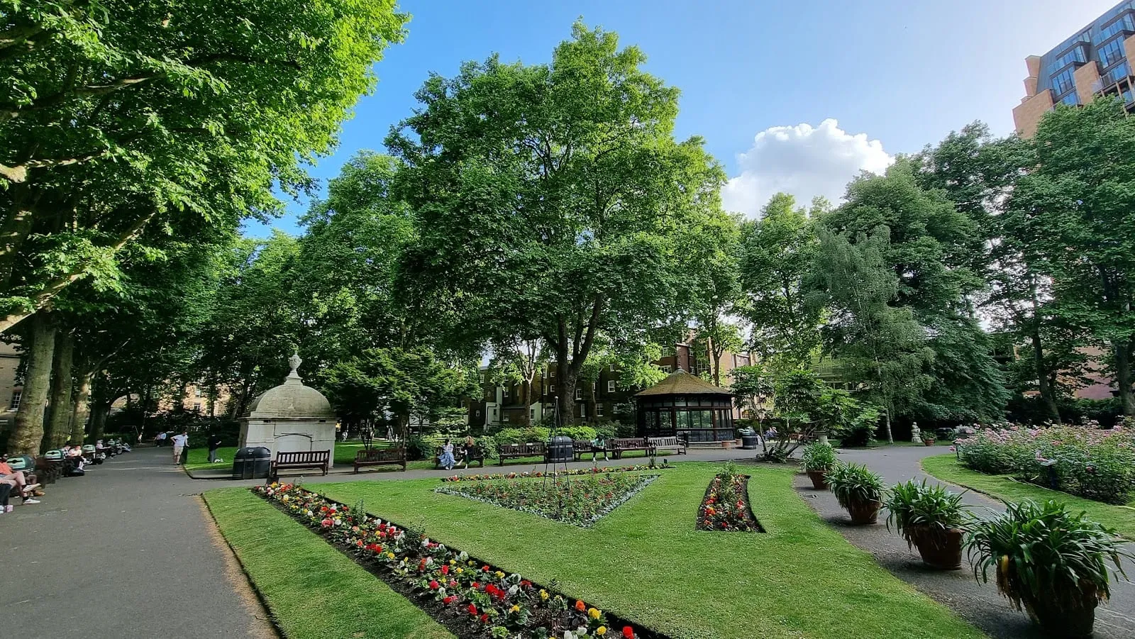 Paddington Street Gardens with a glass-sided bandstand, flower beds and mature plane trees against a blue sky