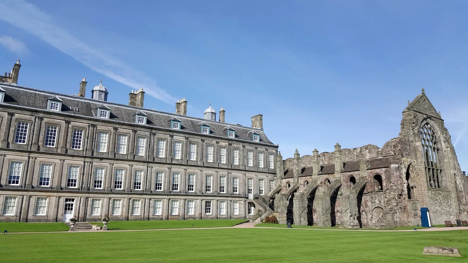 The Palace of Holyroodhouse's grand façade beside the ruined arches and window of the adjoining Holyrood Abbey, under a clear sky