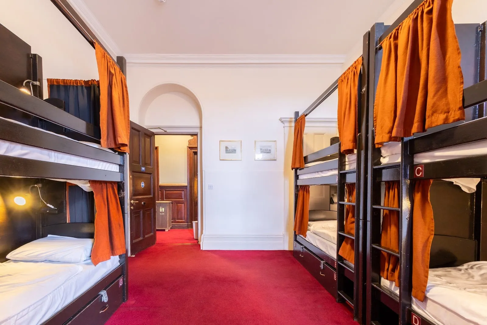 One of Palmers Lodge's large dorms, with black-framed bunk beds, orange privacy curtains and a red carpet running to the corridor