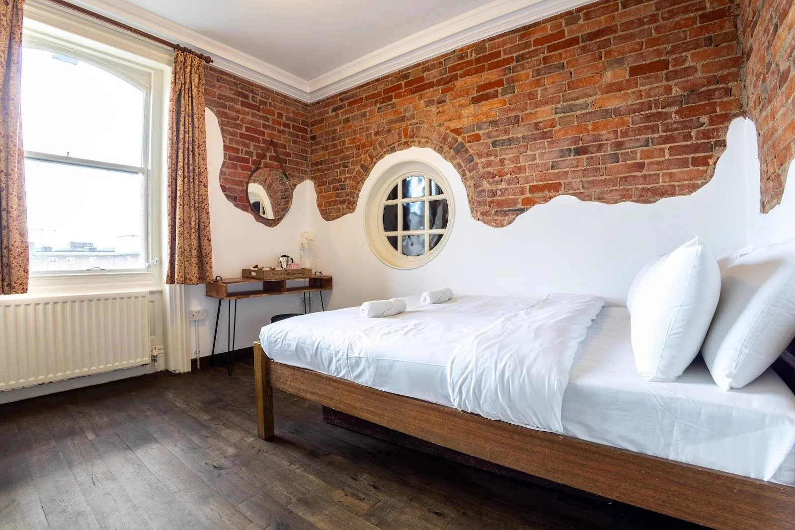 A private double room at Palmers Lodge Swiss Cottage with exposed red brick above white plaster, a round porthole window, a tall sash window, dark floorboards and a plain wooden bed frame