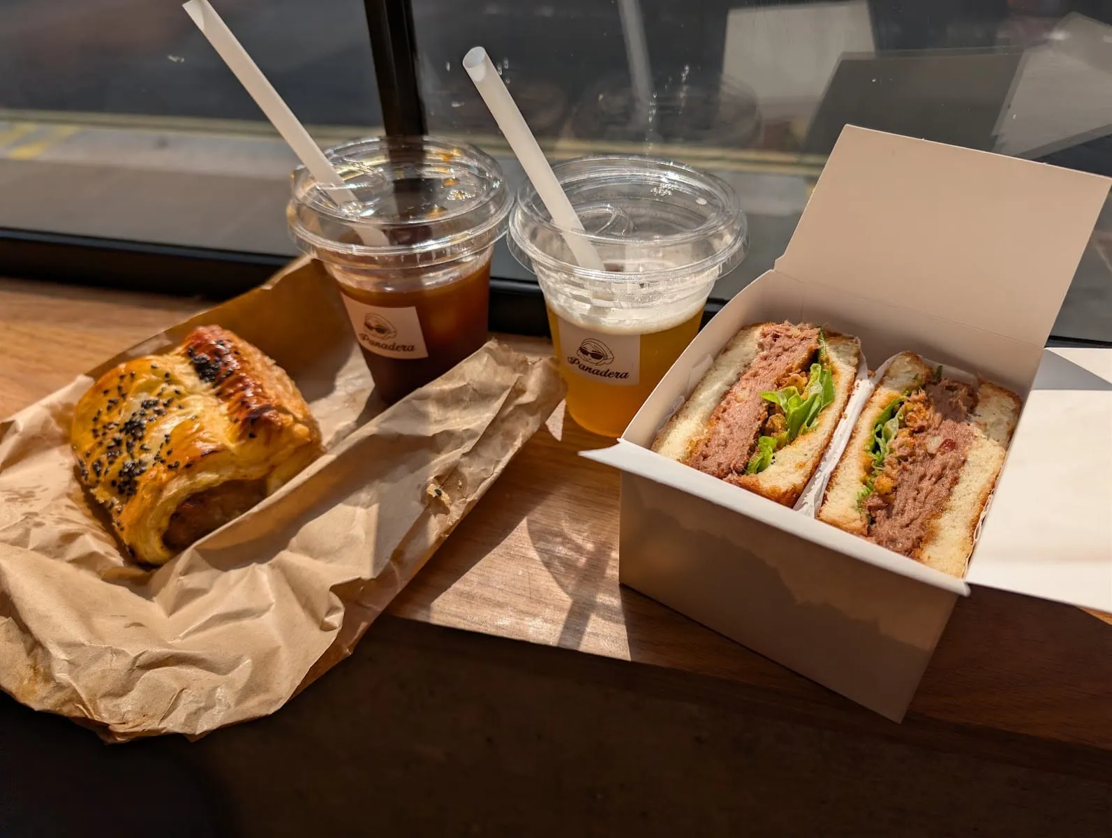A pandesal sando cut in half in an open takeaway box, thick soft bread around a slab of pink meat and lettuce, beside a glazed flaky pastry roll scattered with black sesame seeds and two iced drinks in Panadera-branded cups