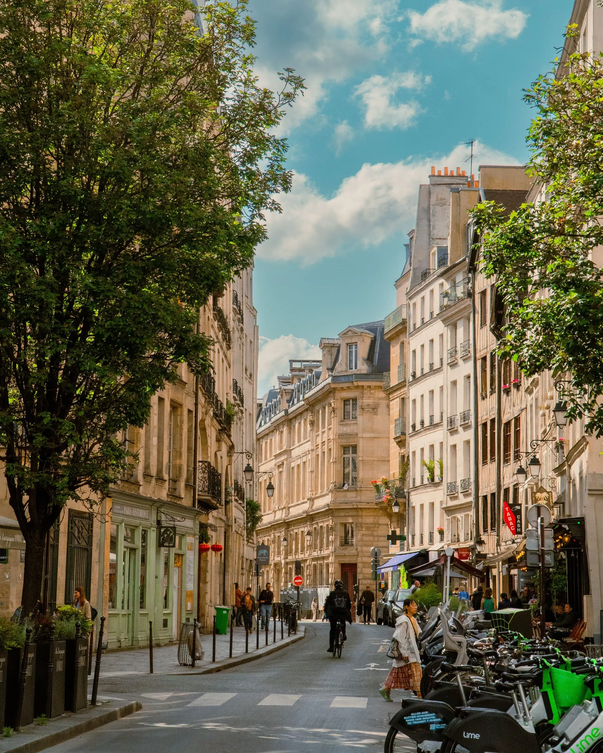 Pale stone apartment buildings along a tree-lined side street in central Paris, with café tables and a row of hire bikes