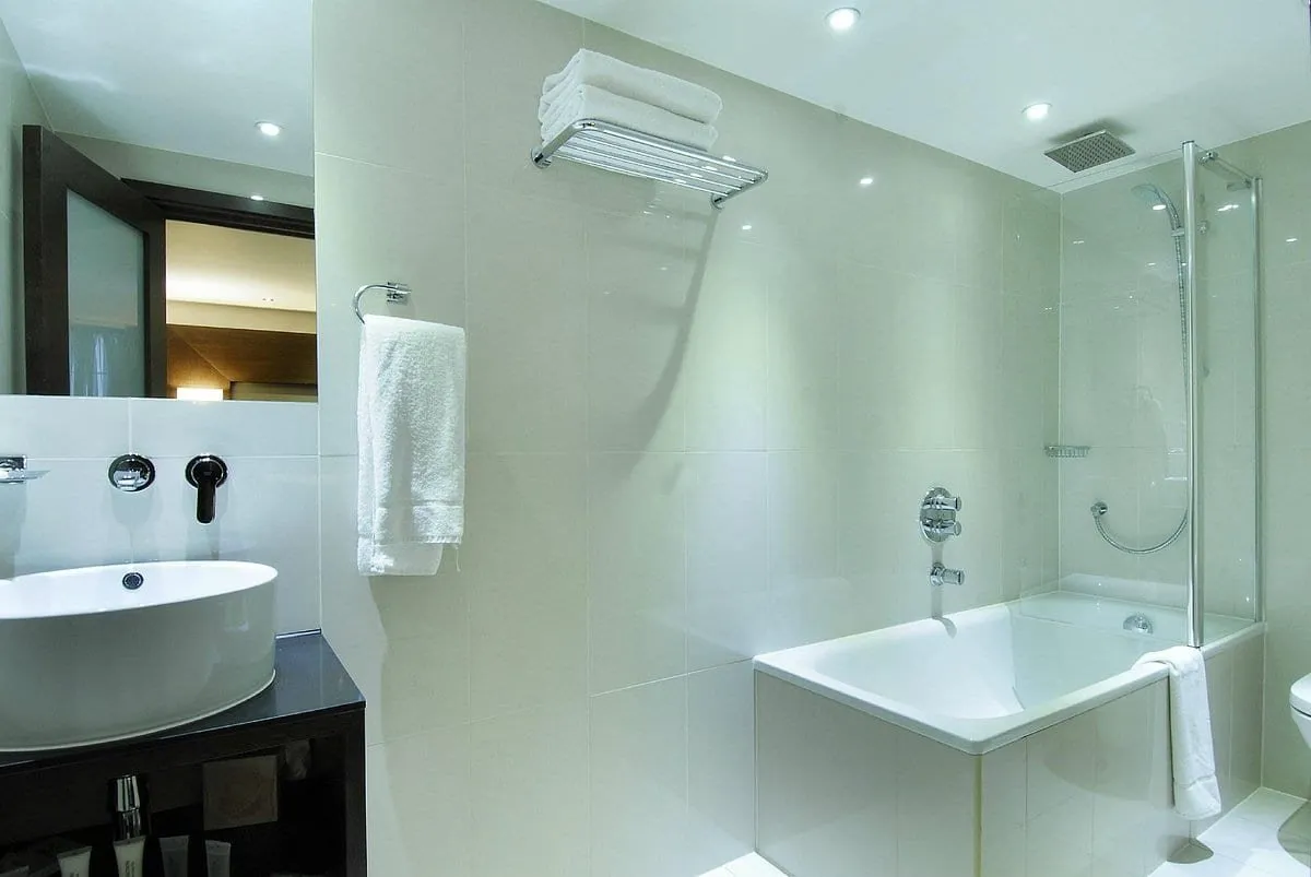 A white-tiled bathroom at Park Avenue Baker Street with a bowl basin on a dark vanity, a towel shelf and a bath with a glass shower screen and a ceiling rain head