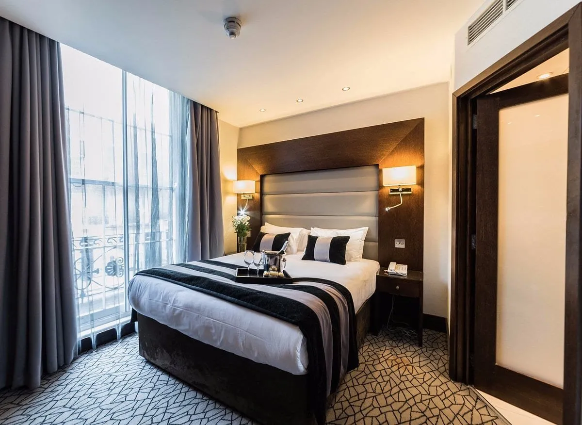 A double room at Park Avenue Baker Street with a grey padded headboard set in a dark wood surround, black and white striped cushions, a patterned carpet and a floor-to-ceiling window with an iron balcony rail