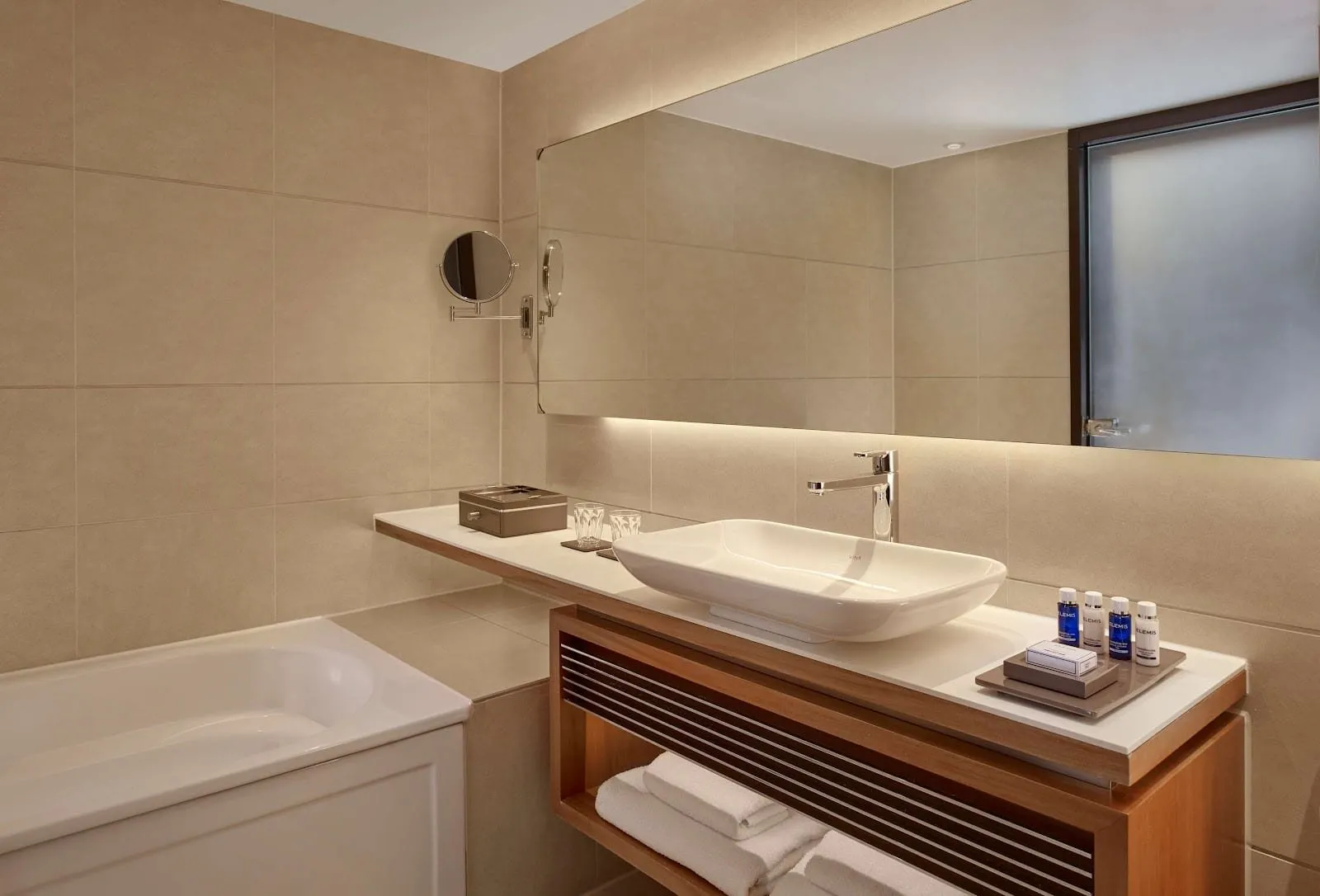 A bathroom at Park Plaza Westminster Bridge with a bath, a rectangular basin on a wooden stand, beige tiles and a long backlit mirror