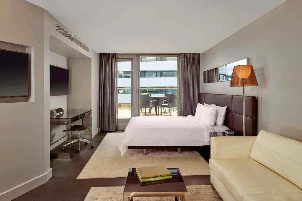 A room at Park Plaza Westminster Bridge with a brown leather headboard, a cream sofa, a glass desk and glass doors onto a terrace with a table and chairs