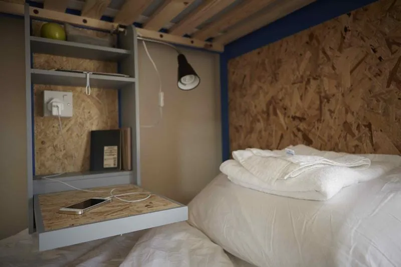 Inside a bunk pod at Park Villa, lined in OSB board, with a reading lamp, a small shelf and a phone charging beside the made-up bed