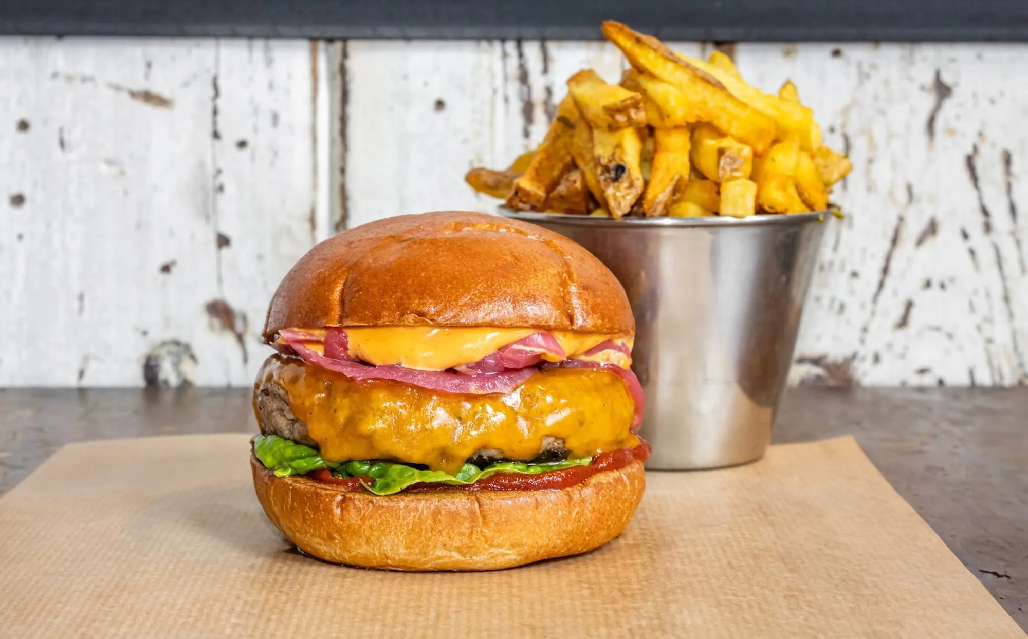 A cheeseburger with a slice of melted cheese, pink pickled onion and lettuce in a glazed bun, next to a metal bucket of thick-cut chips
