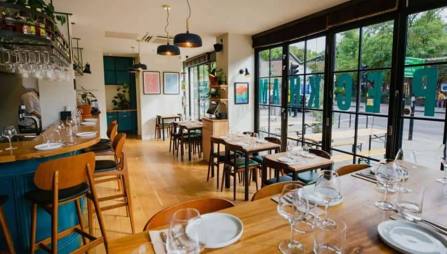 The dining room at Peckham Cellars, light wood tables laid with wine glasses and a teal bar, with PECKHAM lettering across the window onto Queens Road