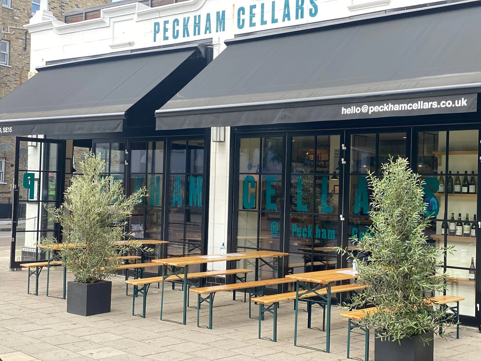 The grey-awninged frontage of Peckham Cellars on Queens Road, with outdoor benches and olive trees