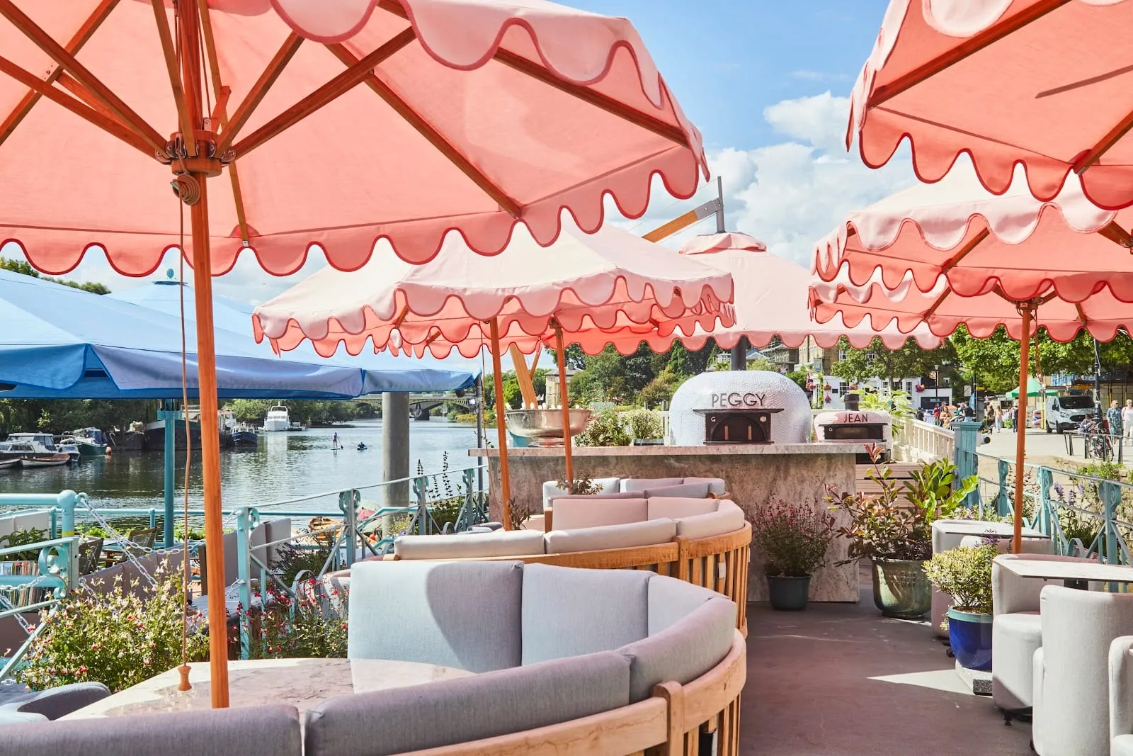 The top deck of the Peggy Jean barge at Richmond: scalloped pink and cream parasols over curved rattan banquettes and marble tables, two domed wood-fired ovens lettered "Peggy" and "Jean", and the Thames with moored boats and Richmond Bridge behind
