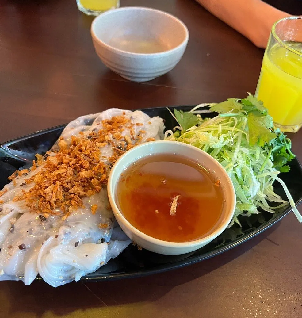 Steamed rice rolls topped with crisp fried shallots on a black oval plate, with a bowl of dipping sauce and a pile of shredded herbs beside them