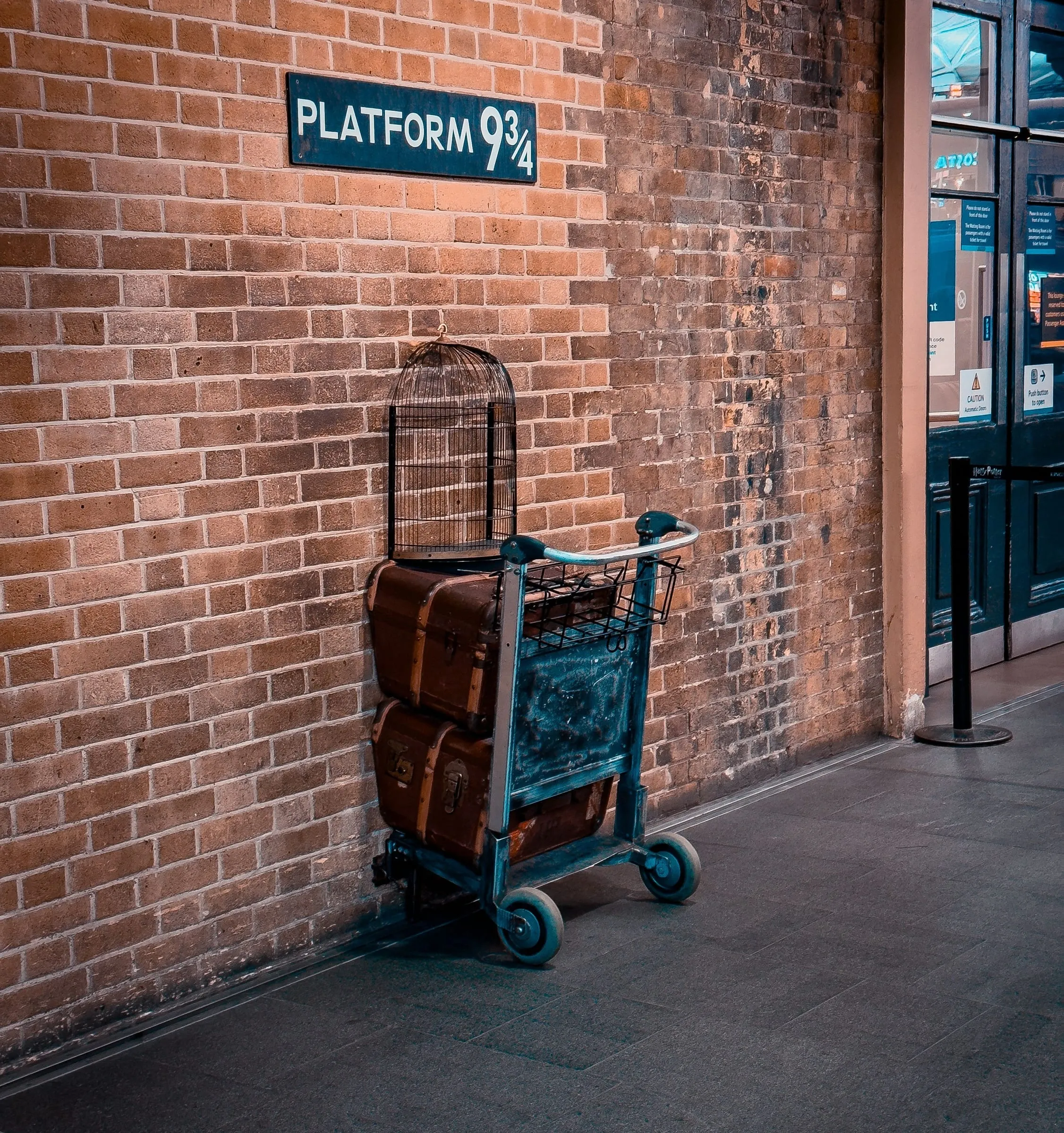A luggage trolley loaded with trunks and an owl cage, standing against the brick wall beneath the Platform 9¾ sign at King's Cross