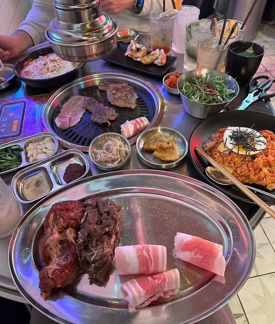 A table at Pochawa Grill under pink and blue neon, an extractor hood over the grill, a steel platter of raw marinated beef and rolled pork belly, and a bowl of kimchi fried rice topped with a fried egg