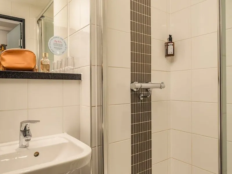 A bathroom at Point A Hotel London Canary Wharf with a shower cubicle behind a glass door, white tiles with a grey mosaic strip, and a small basin under a mirror shelf
