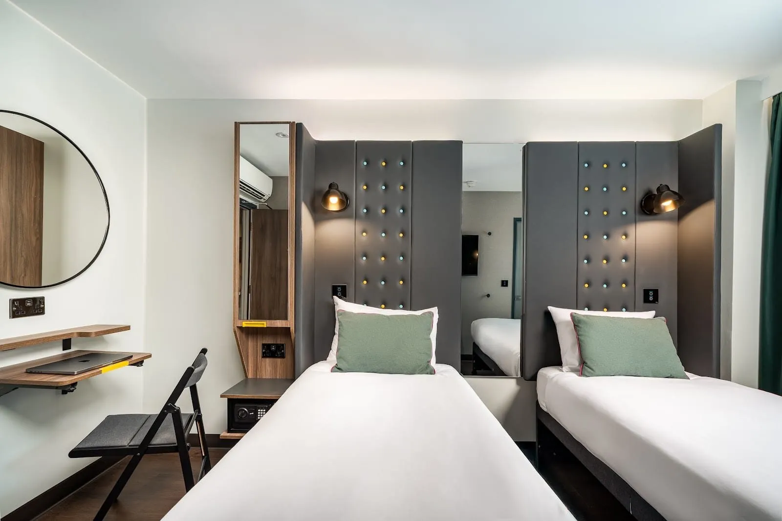 A twin room at Point A Hotel London Canary Wharf with two single beds against grey padded headboards studded with blue and yellow buttons, a fold-down desk and chair, and a round mirror
