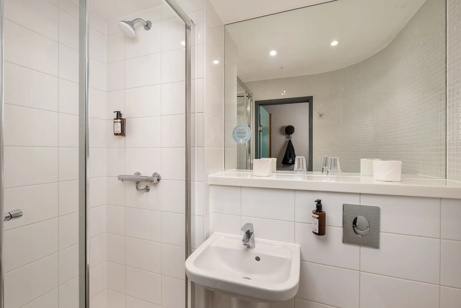 A compact white-tiled shower room at Point A Hotel London Paddington with a glass shower cubicle, a small basin and a mirror over a narrow shelf