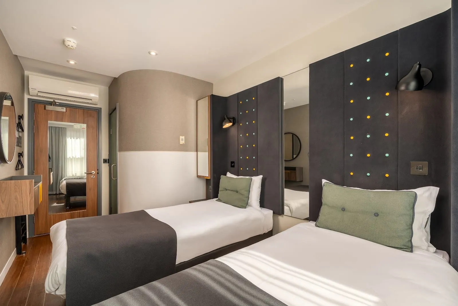 A twin room at Point A Hotel London Paddington with two single beds against tall charcoal headboards studded with yellow and mint dots, a curved wall, wall-mounted air conditioning and a window reflected in the door mirror