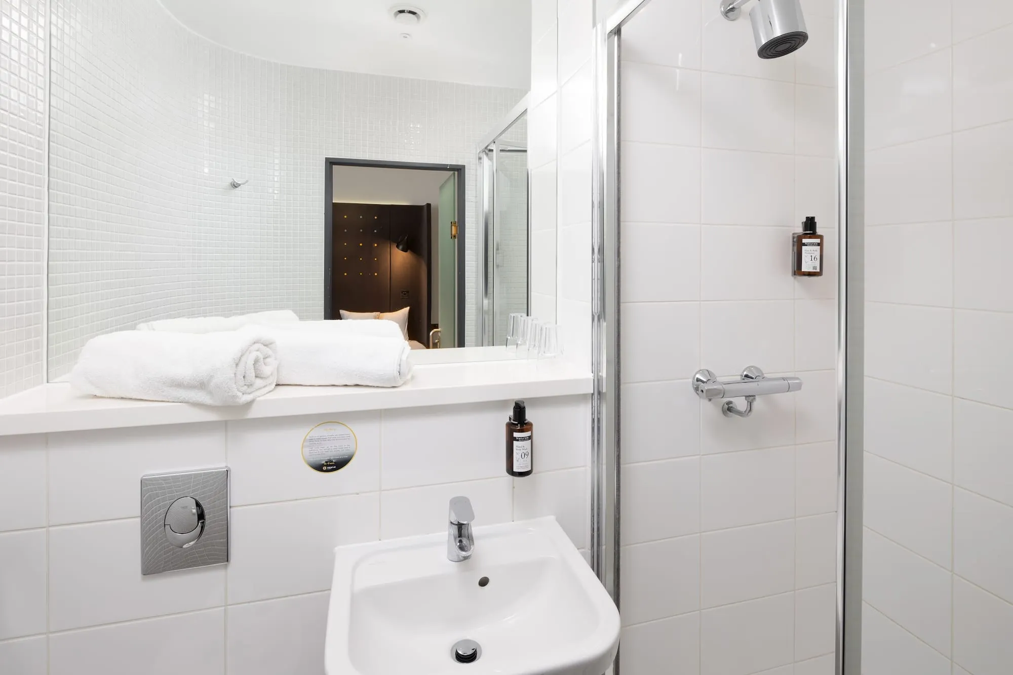 The ensuite bathroom of a Point A Hotel King's Cross room, with a glass-screened shower, a wall-mounted sink and rolled towels on the counter