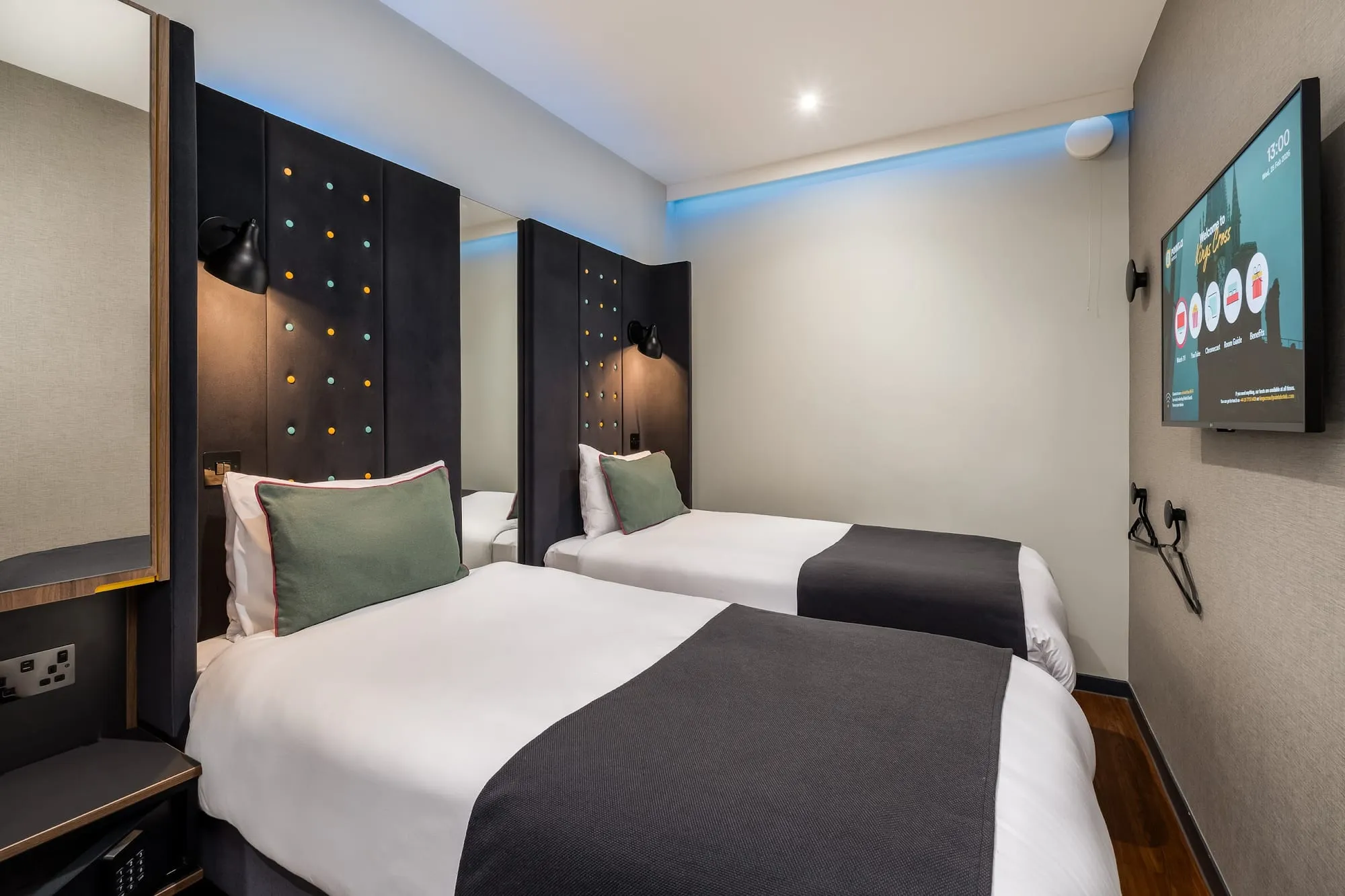 A twin room at Point A Hotel King's Cross, with a button-studded black headboard, blue cove lighting along the ceiling, and a wall-mounted TV showing a King's Cross welcome screen