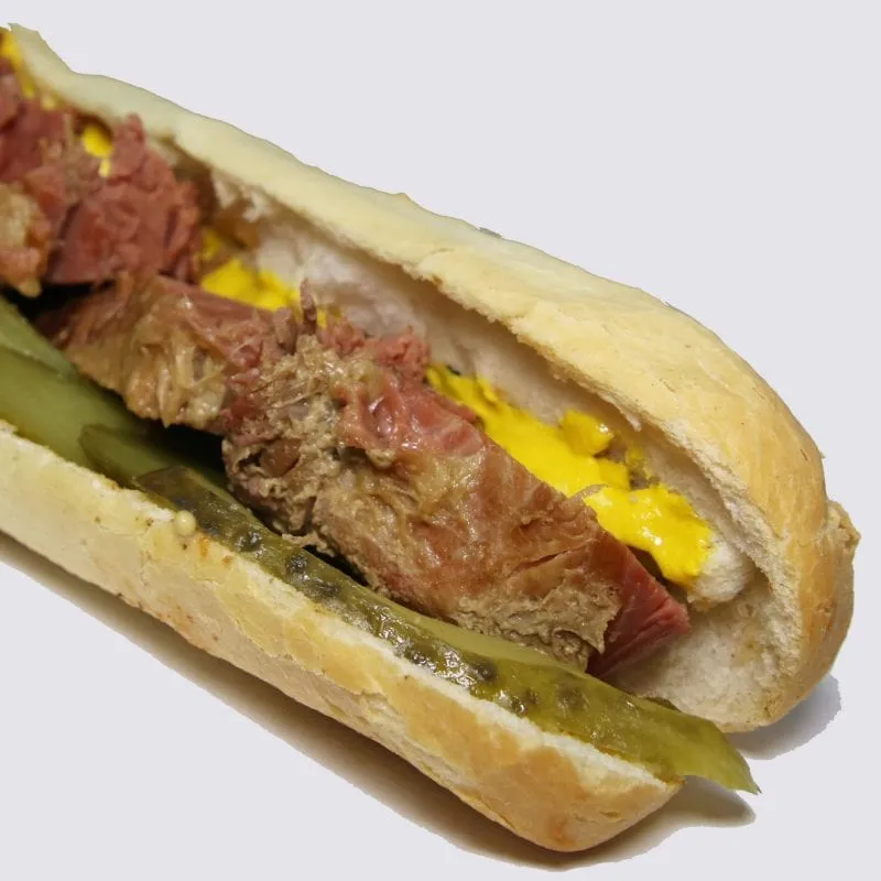 A salt beef baguette cut open, filled with hand-carved beef, yellow mustard and a whole gherkin laid along the length of it