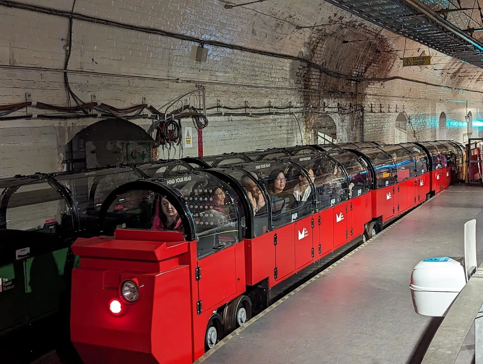 A red miniature train with glass-domed carriages carrying passengers through a brick-lined tunnel at the Postal Museum's Mail Rail